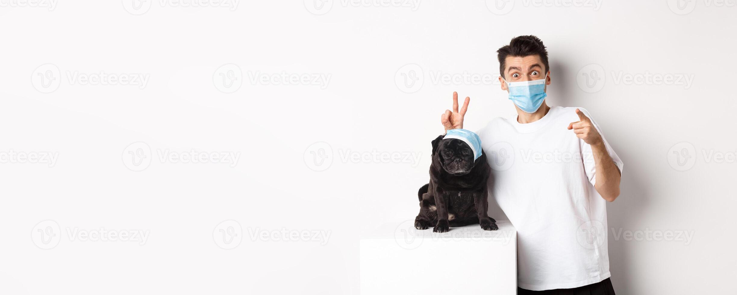 Covid-19, animals and quarantine concept. Happy dog owner and cute pug wearing medical masks, man pointing finger at camera and making funny bunny ears on pet, white background photo