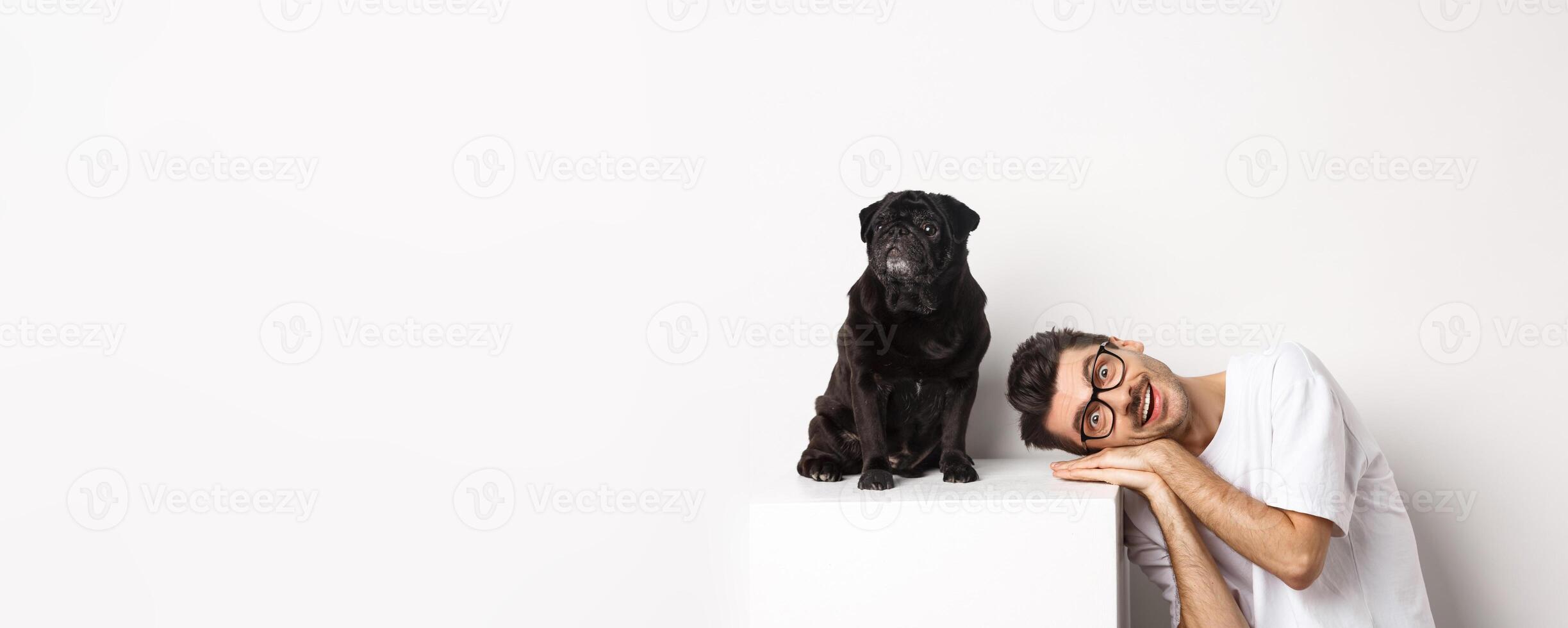 Handsome young man lay head near cute black dog, smiling and looking at camera, white background photo