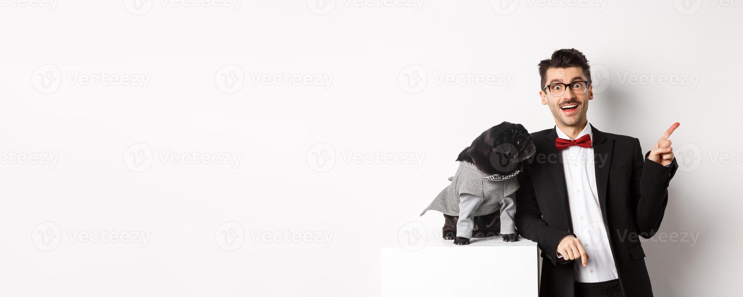 Animals, party and celebration concept. Happy and amazed young man and cute pug in costume standing over white background, dog owner pointing right photo