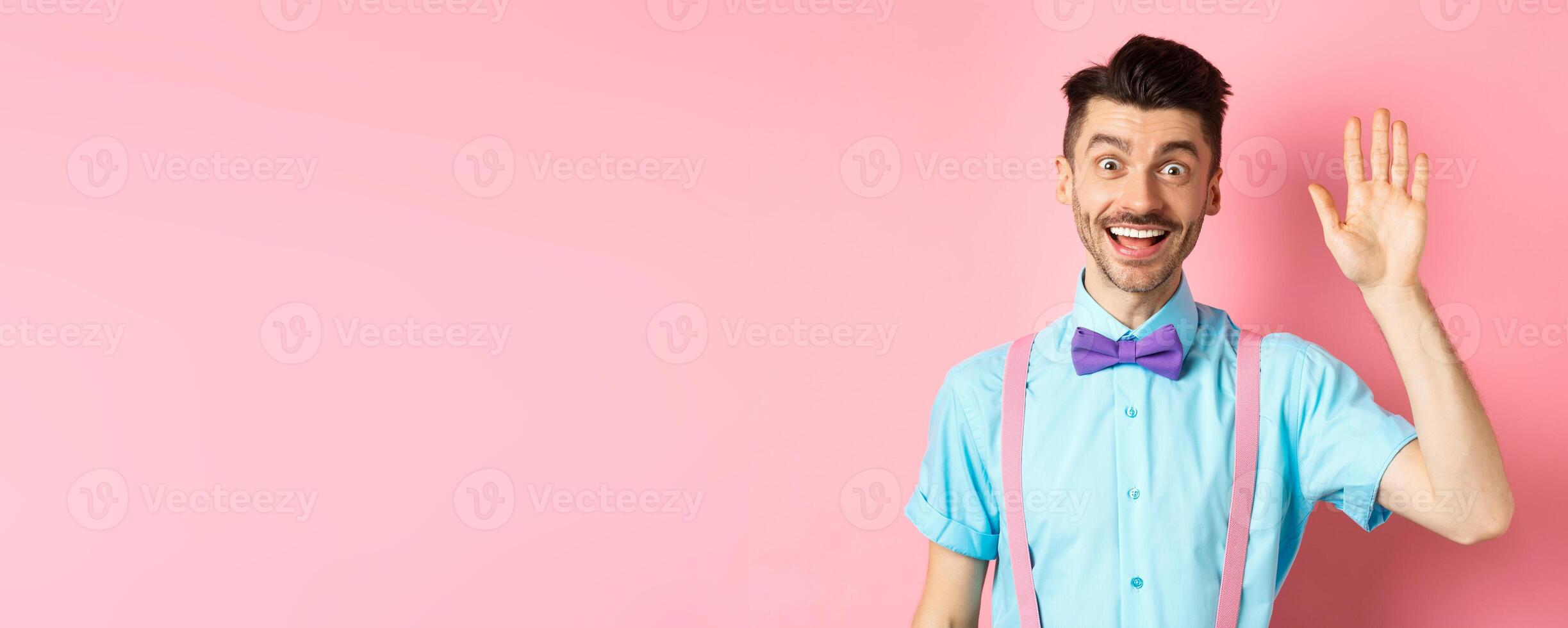 Friendly smiling man in funny bow-tie saying hello, waving hand to greet you, make hi gesture and looking happy yo see you, standing over pink background photo