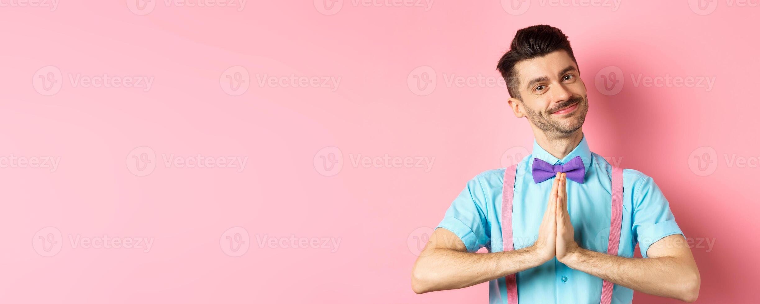 Smiling young guy in bow-tie say thank you, looking with gratitude and holding hands in namaste gesture, standing over pink background photo