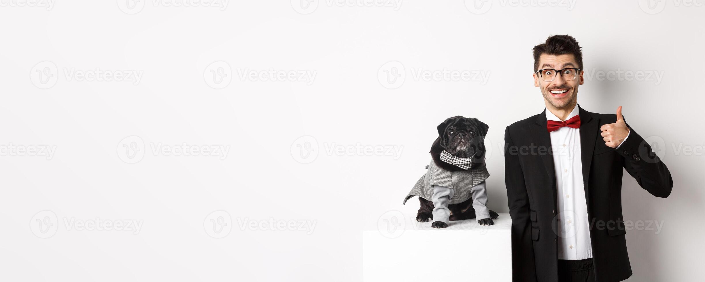 Animals, party and celebration concept. Handsome young man in suit and cute black pug in costume staring at camera, owner showing thumb up in approval and praise, white background photo