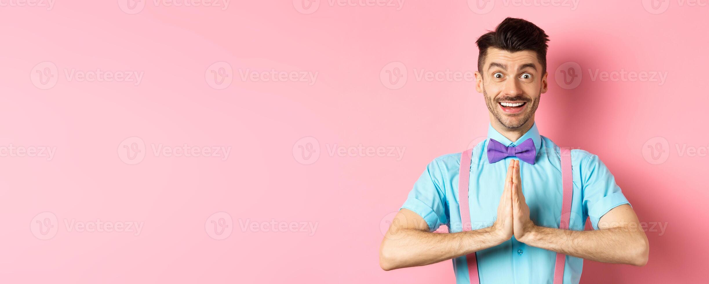 Image of smiling young man asking please, looking grateful at camera, Guy in bow-tie say thank you, standing over pink background photo
