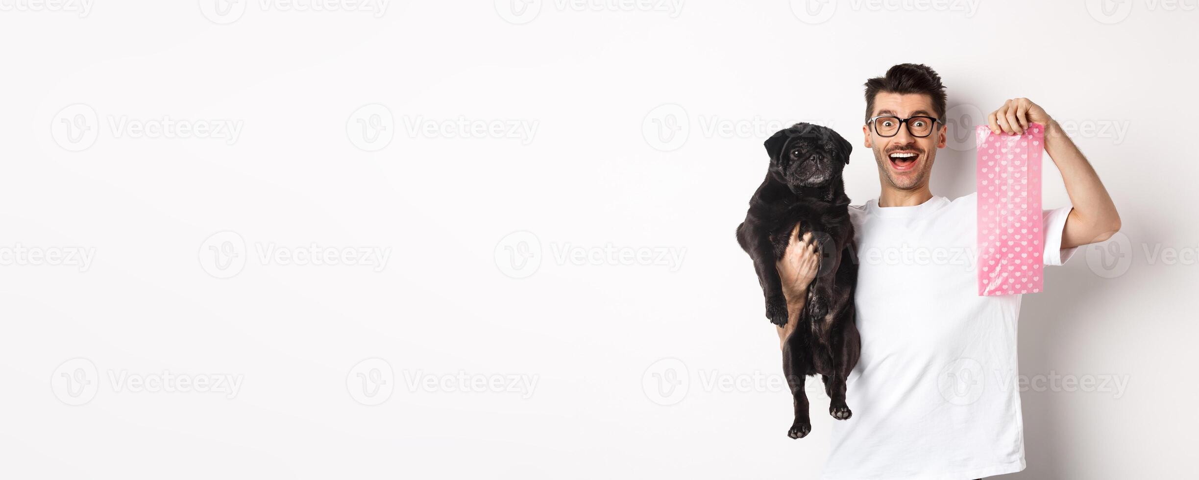 Image of hipster guy pet owner, holding cute black pug and dog poop bag, standing over white background photo