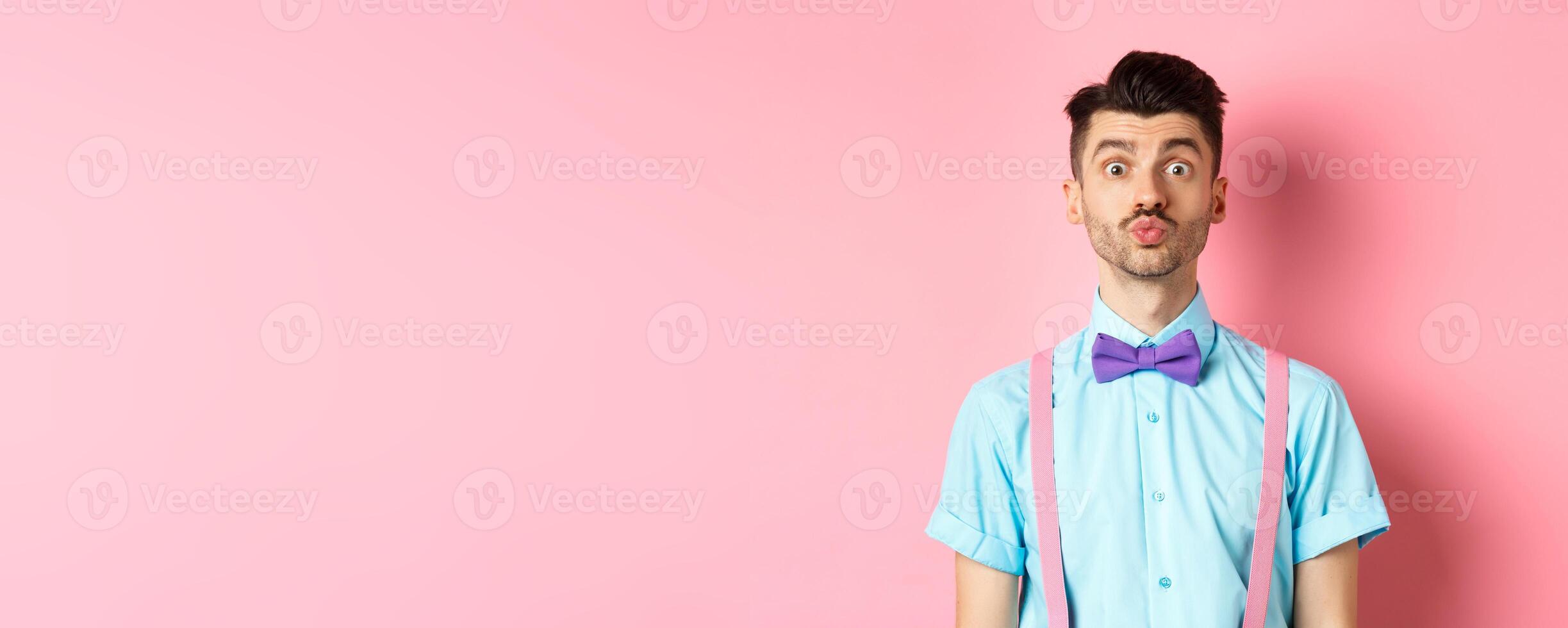 Funny young man waiting for kiss with puckered lips and silly face, standing on romantic pink background. Concept of love and Valentines day photo