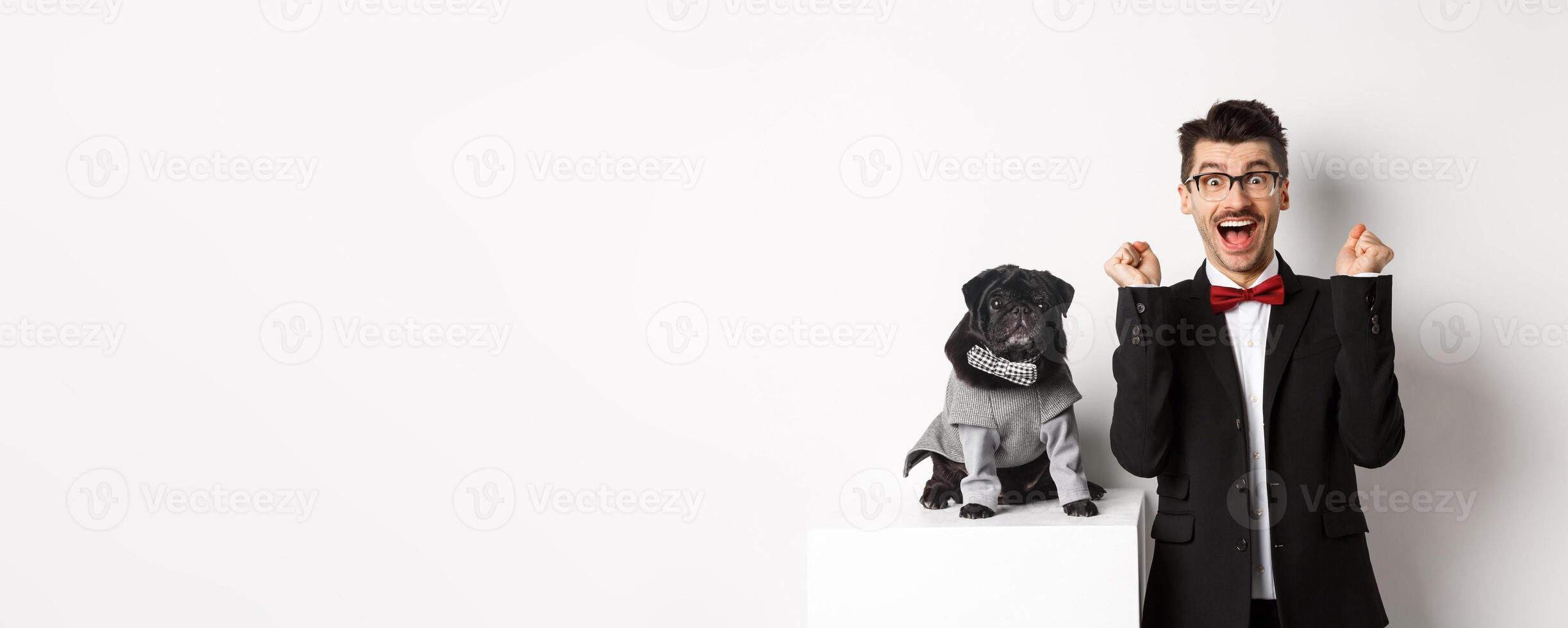 Animals, party and celebration concept. Happy young man in suit and puppy in pet cosume standing over white background, dog owner rejoicing and triumphing photo