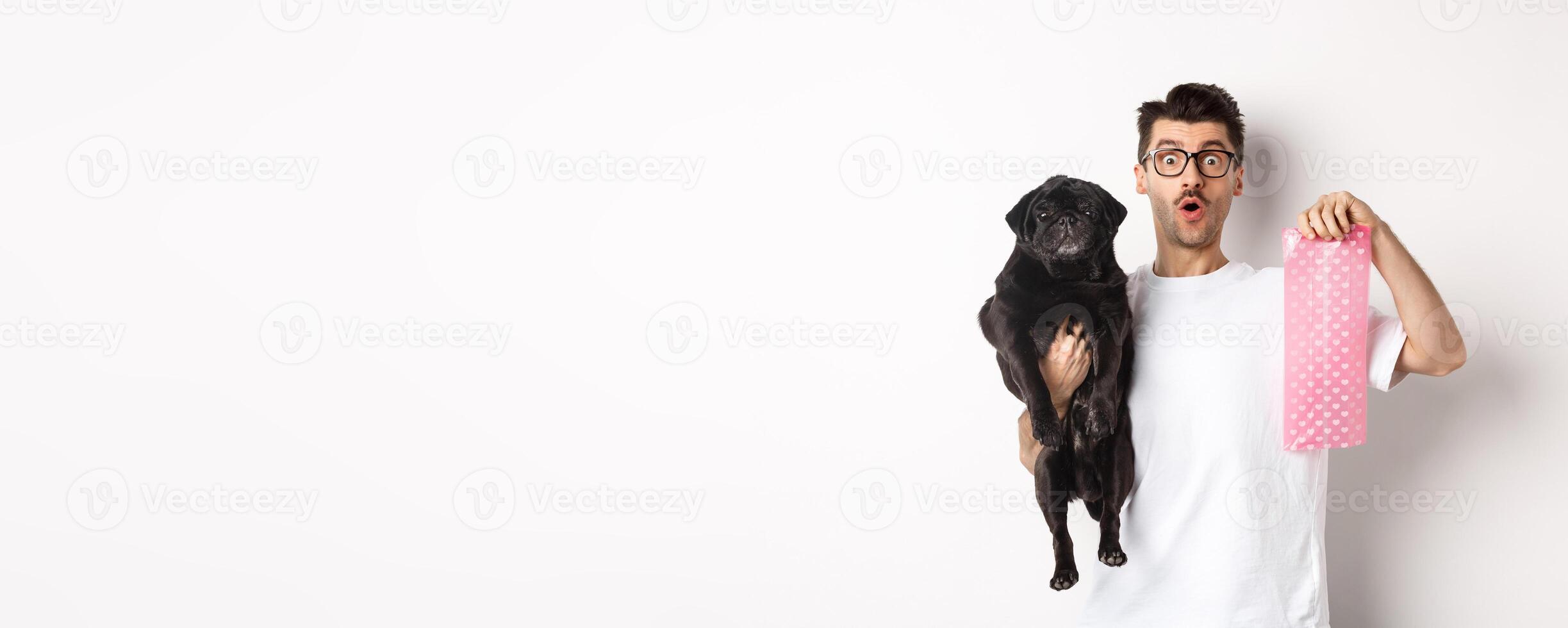 Image of hipster guy pet owner, holding cute black pug and dog poop bag, standing over white background photo