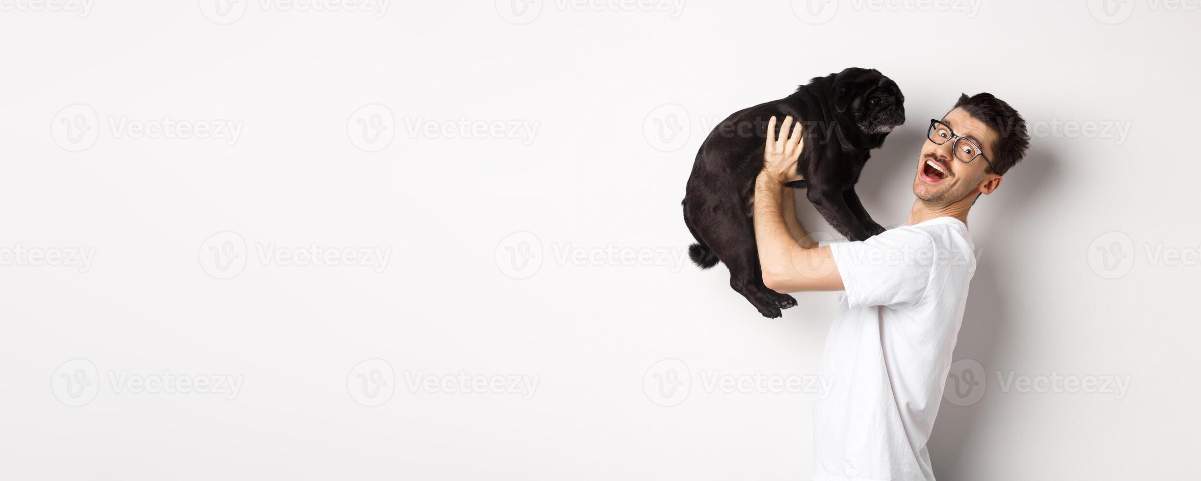 Image of handsome young man loving his pug. Dog owner holding puppy and smiling happy at camera, standing over white background photo