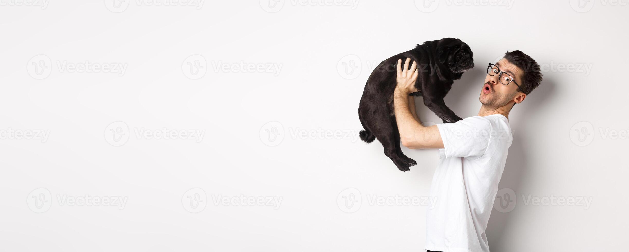 Image of handsome young man loving his pug. Dog owner holding puppy and smiling happy at camera, standing over white background photo
