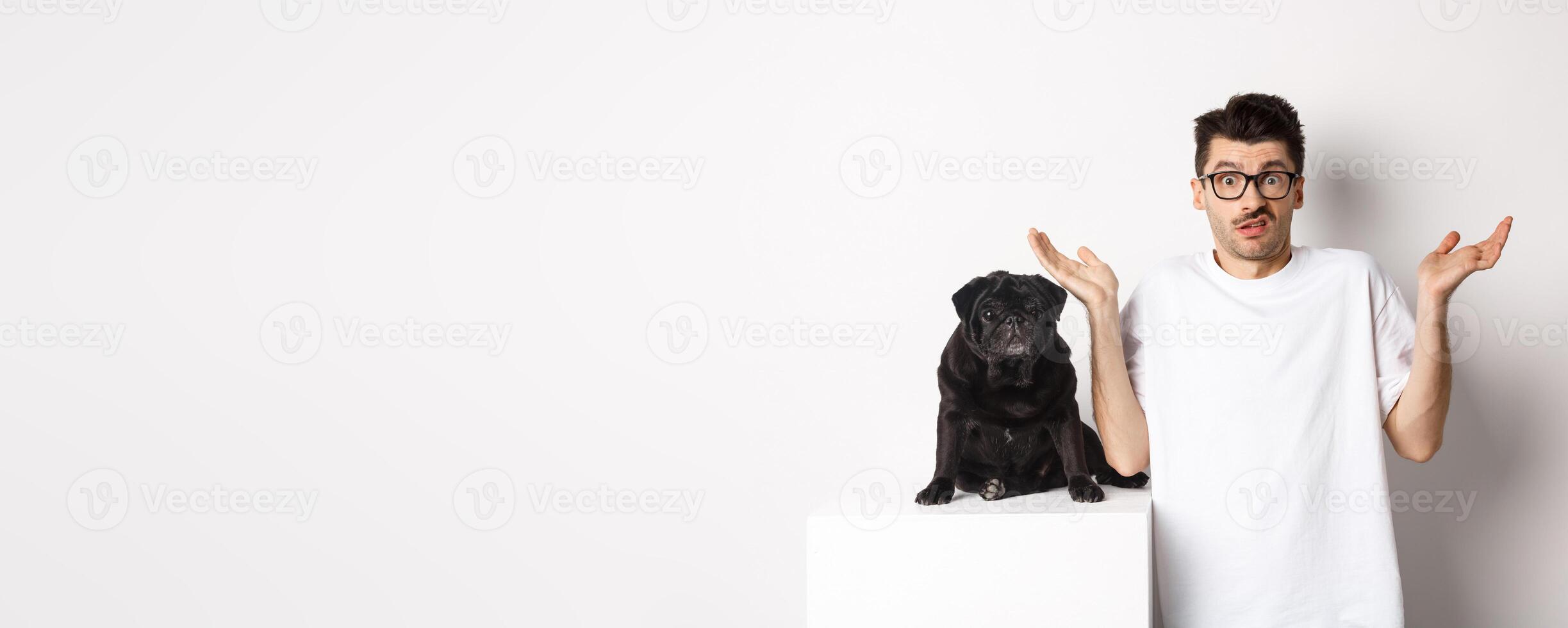 Image of confused man in glasses raising hands and shrugging complicated, standing near black pug dog over white background photo
