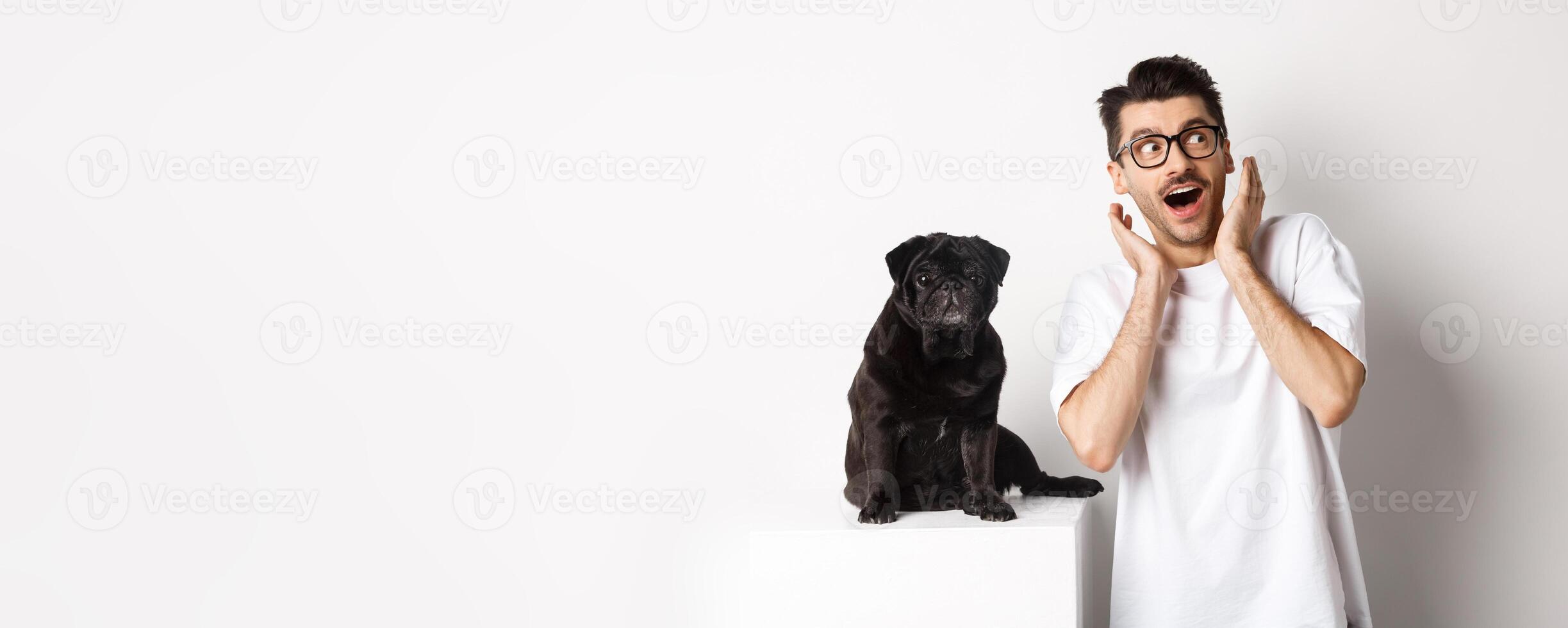 Amazed young man standing with cute black puppy, staring at upper right corner surprised and excited, standing near pug over white background photo