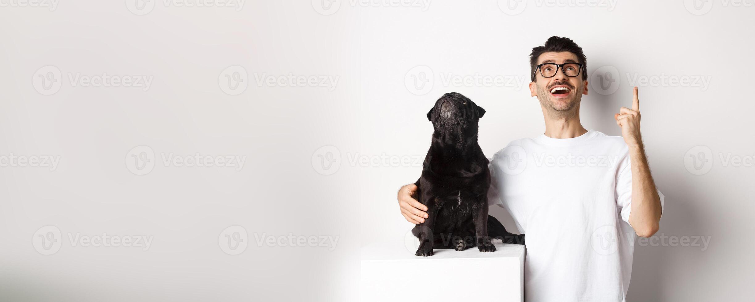 Image of happy young man and dog both looking up at promo, owner pointing finger at top, pug staring at loog, standing over white background photo
