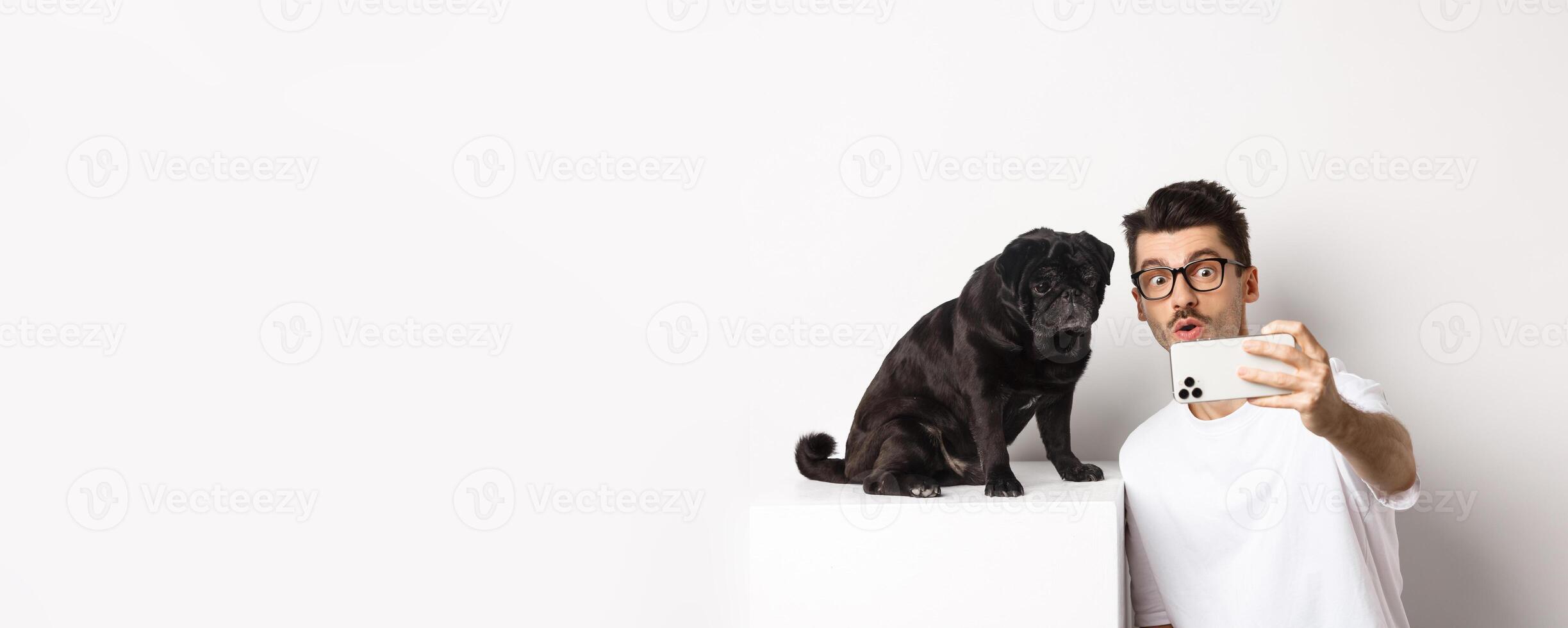 Image of handsome young man taking selfie with cute black dog on smartphone, posing with pug over white background photo