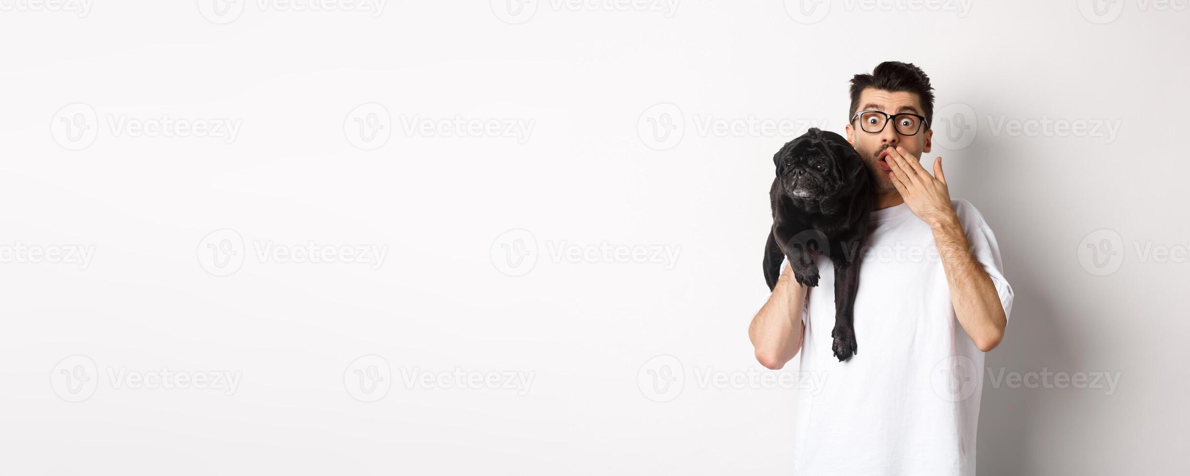 Shocked dog owner staring at camera, holding puppy on shoulder and gasping amazed. Handsome young man carry pug and reacting to promo advertisement, white background photo