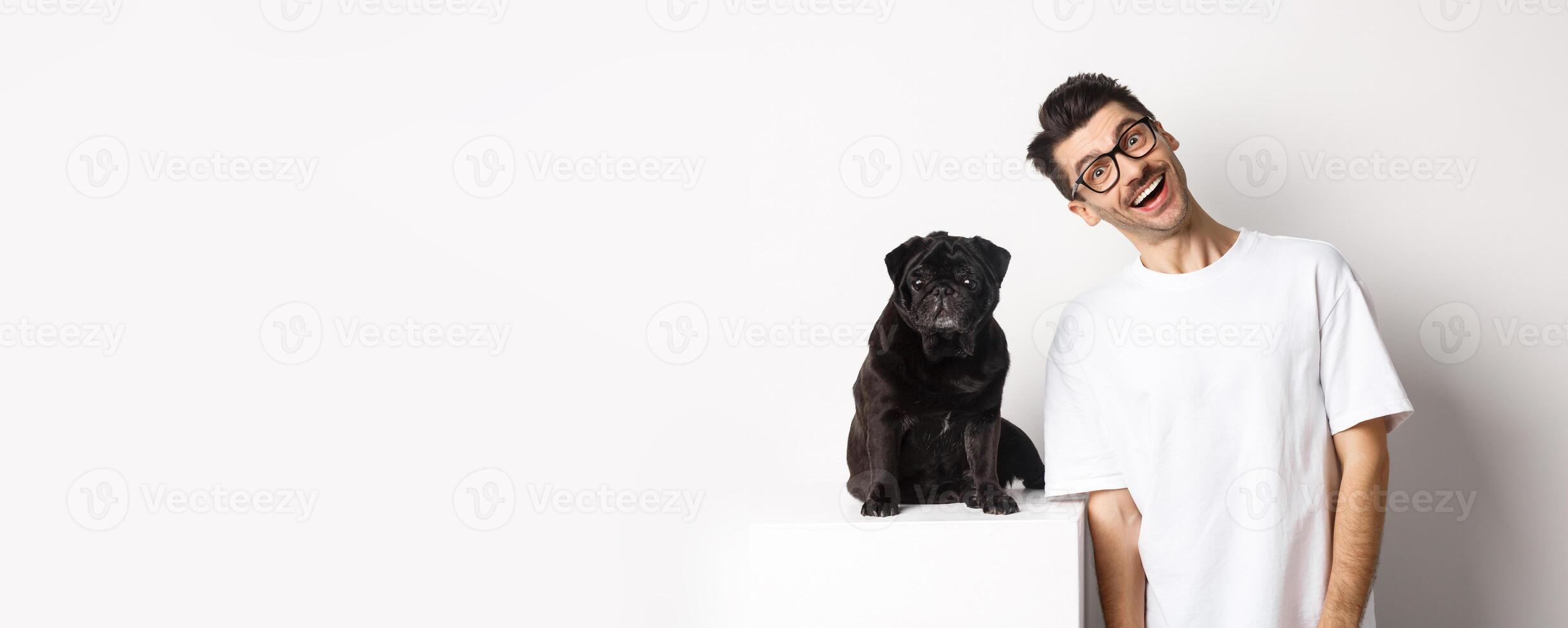 Image of handsome young man standing near cute black pug and smiling. Dog owner spending time with his pet, staring at camera happy, white background photo
