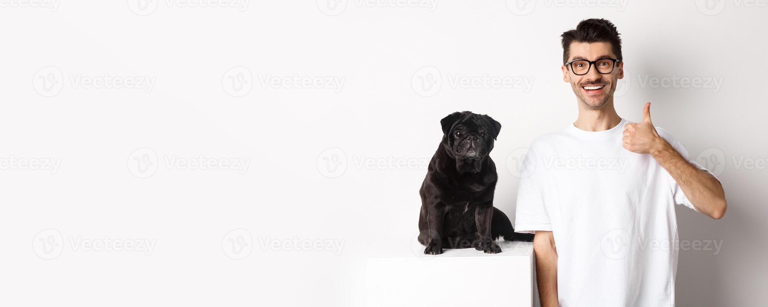 Image of dog owner and cute black pug looking at camera, man showing thumb-up in approval, recommending something, standing over white background photo