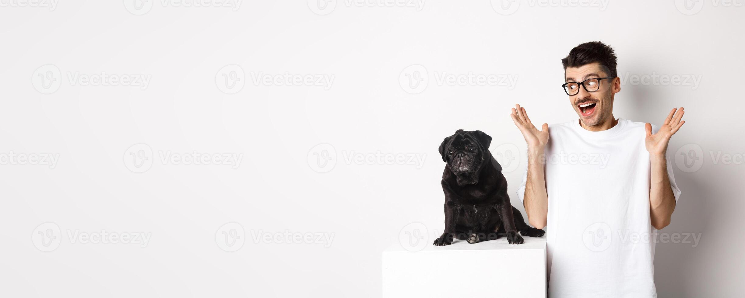 Image of cheerful young man rejoicing, looking at cute black dog pug and smiling, standing over white background photo