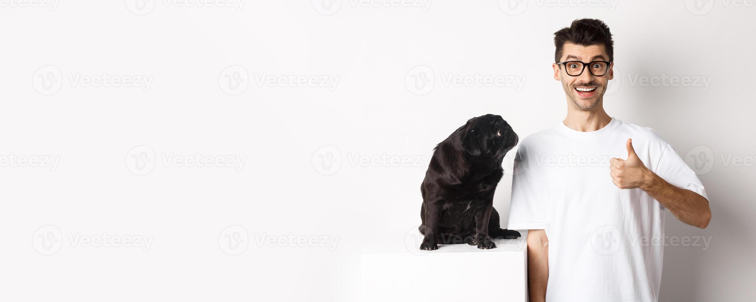Cheerful handsome guy standing near cute black pug and showing thumb-up. Pet owner approve and recommend product for dogs, white background photo