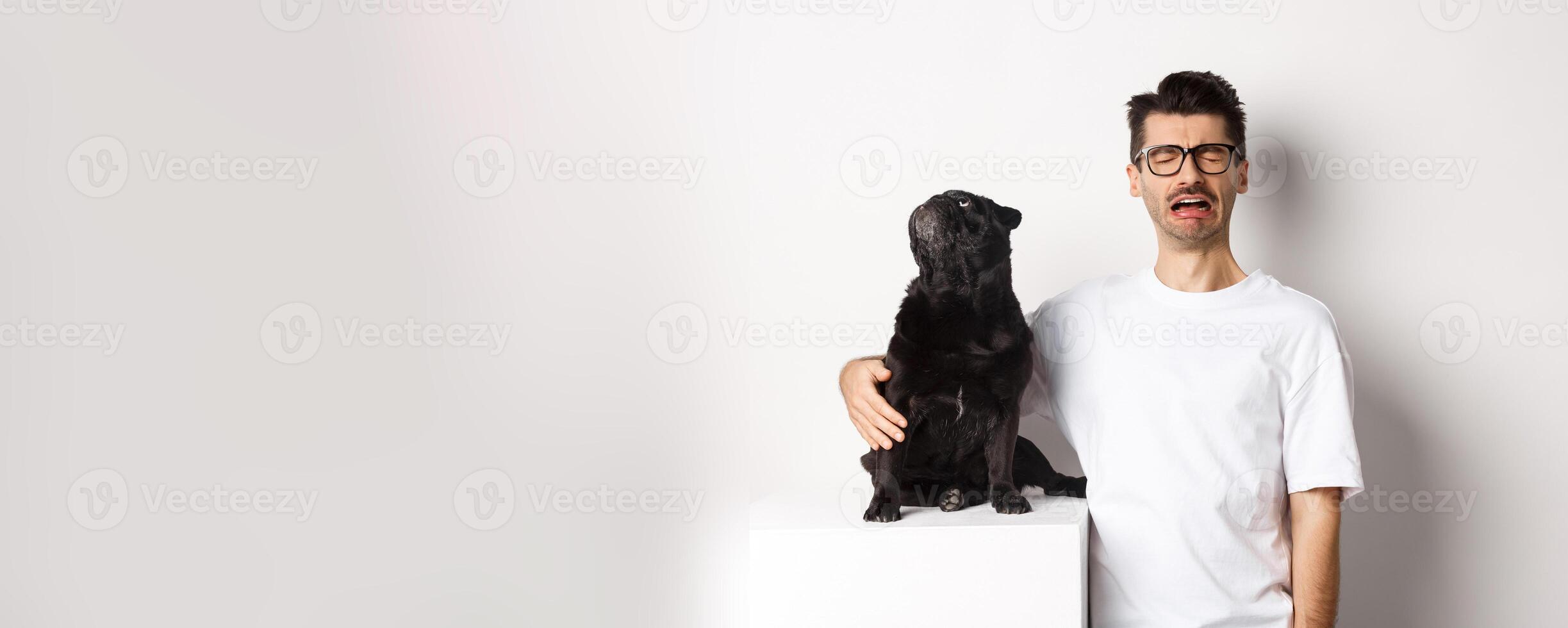 Image of sad crying man hugging his cute black pug, dog looking curious at upper left corner promo logo, standing over white background photo