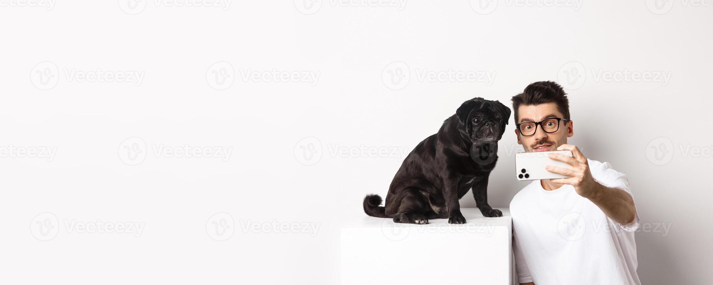 Image of handsome young man taking selfie with cute black dog on smartphone, posing with pug over white background photo