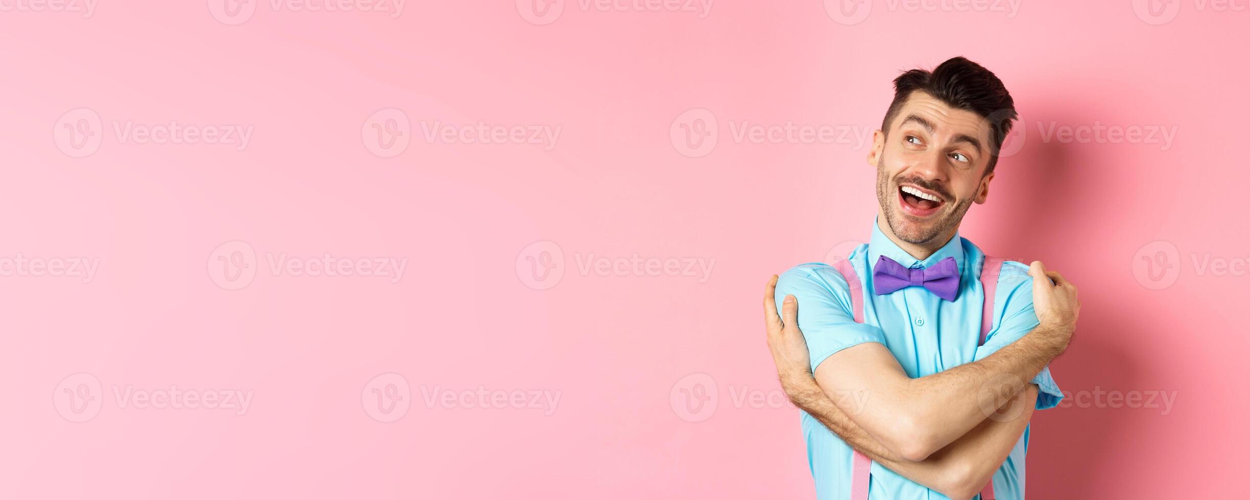 Happy and positive guy in bow-tie, hugging himself and looking left with cheerful smile, standing over pink background photo