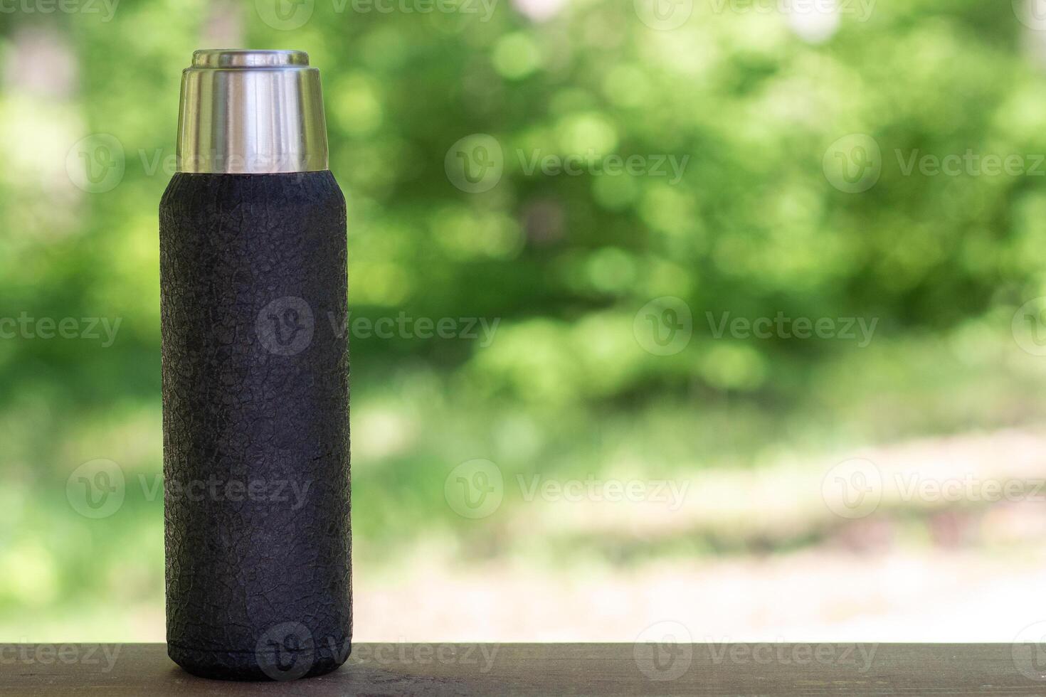 A black thermos with brewed tea or coffee stands on a wooden board with a copyspace. Thermos with hot drink photo