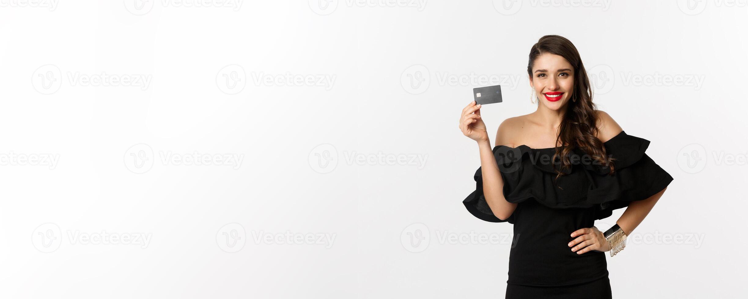 Beauty and shopping concept. Stylish pretty woman with red lips, black dress, smiling happy and showing credit card, standing over white background photo