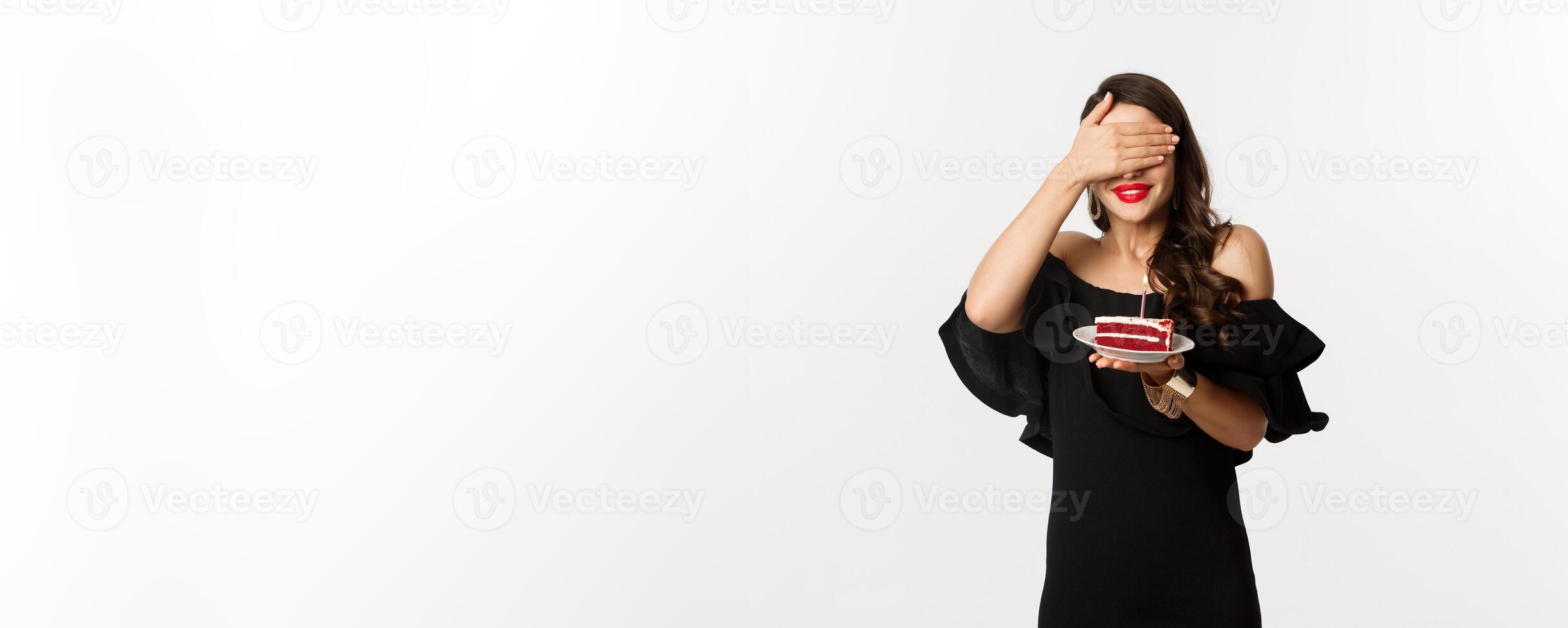 Celebration and party concept. Happy birthday girl in black dress, red lipstick, close eyes and making wish on b-day cake, standing over white background photo