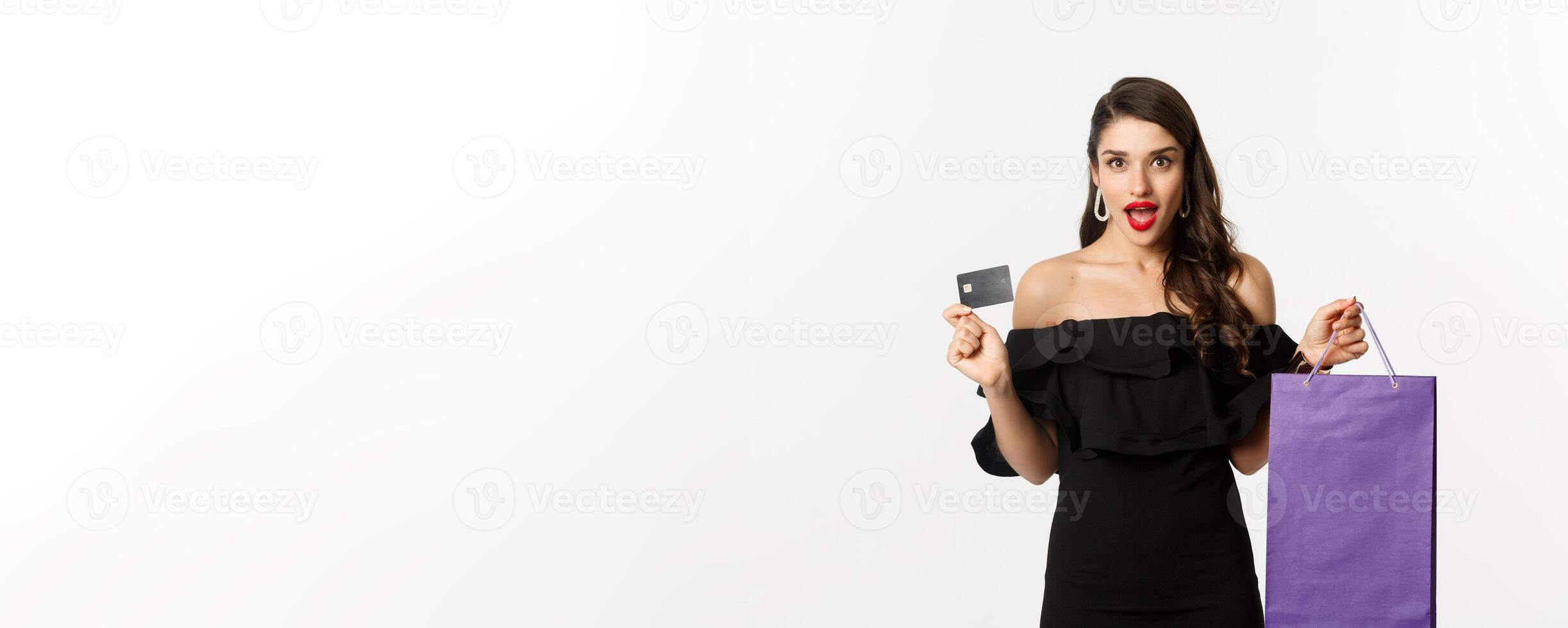 Stylish young woman in black dress going shopping, holding bag and credit card, smiling pleased, standing over white background photo