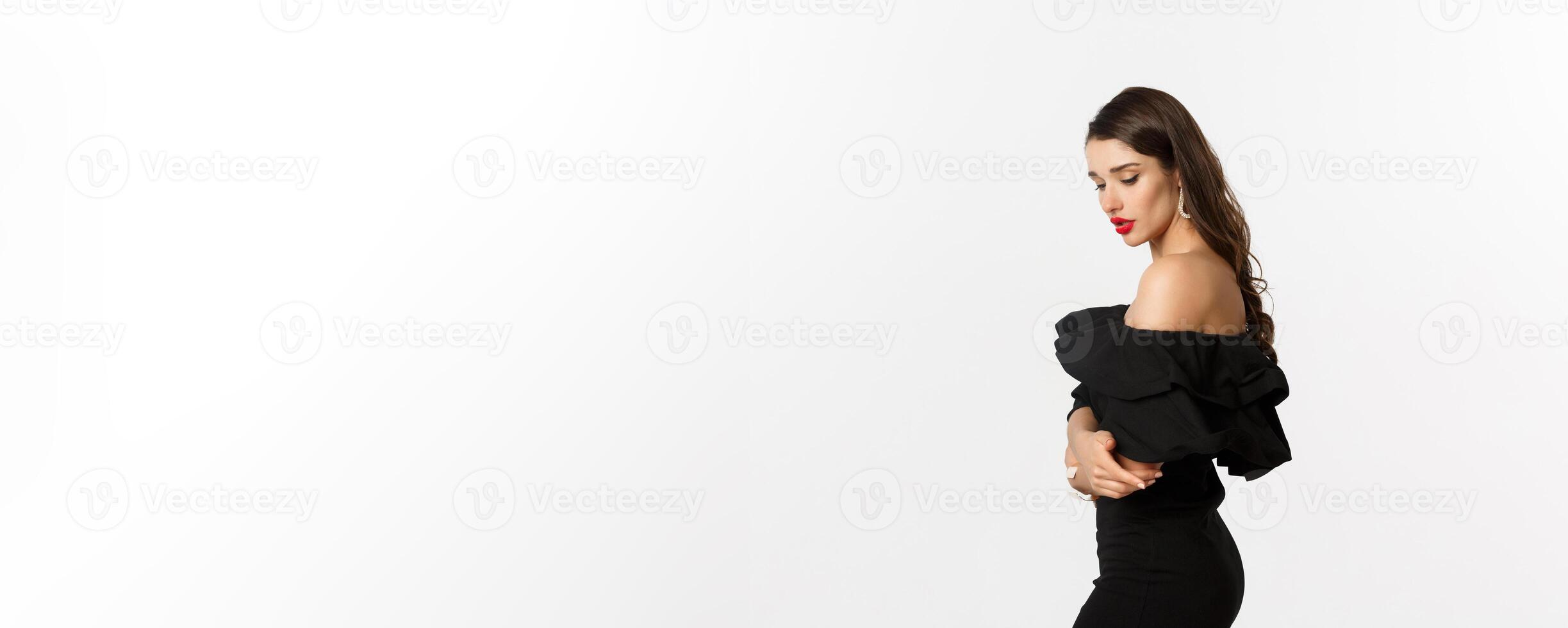 Profile of sensual brunette woman in black elegant dress, looking down, standing over white background photo