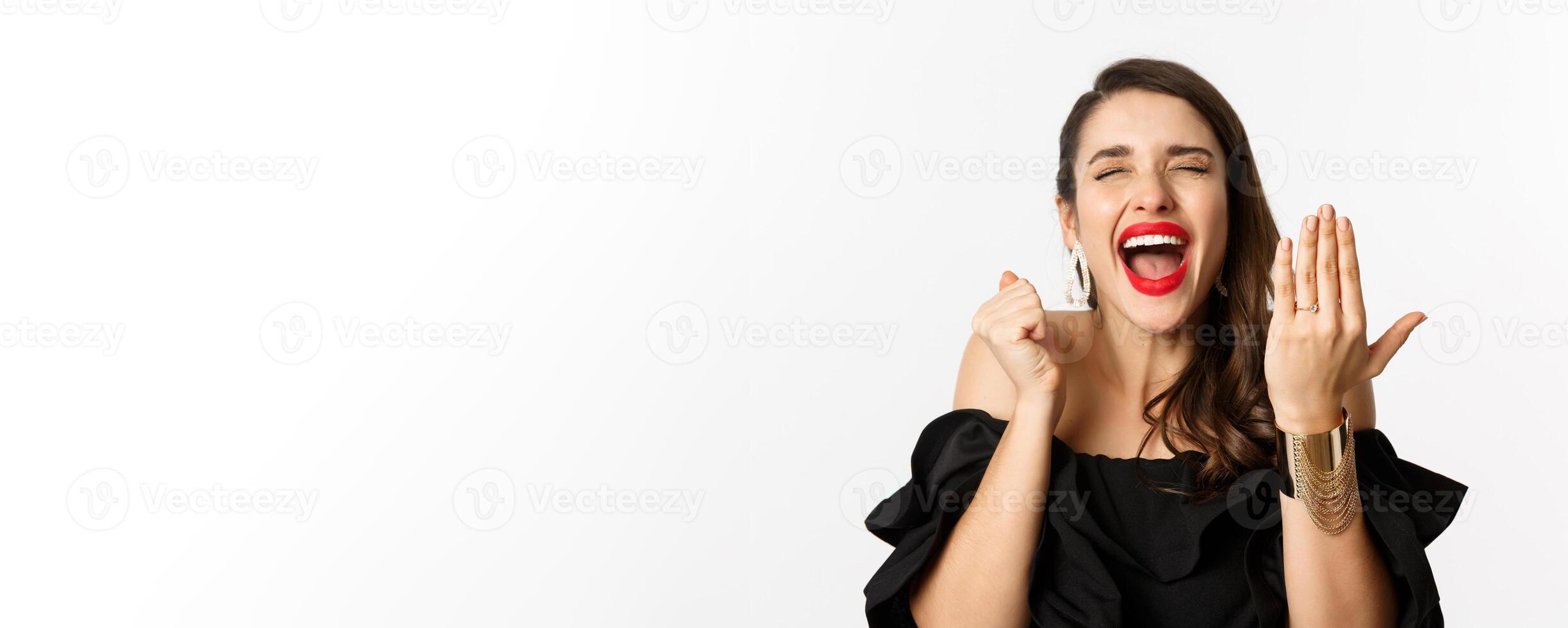 Fashion and beauty concept. Excited and happy woman said yes, showing hand with engagement ring and shouting joyful, become bride, standing over white background photo
