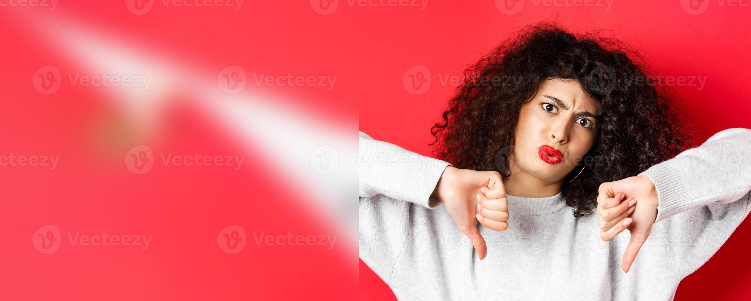 Close-up of disappointed young woman with curly hairstyle, showing thumbs down and frowning, condemn something bad, standing displeased on red background photo