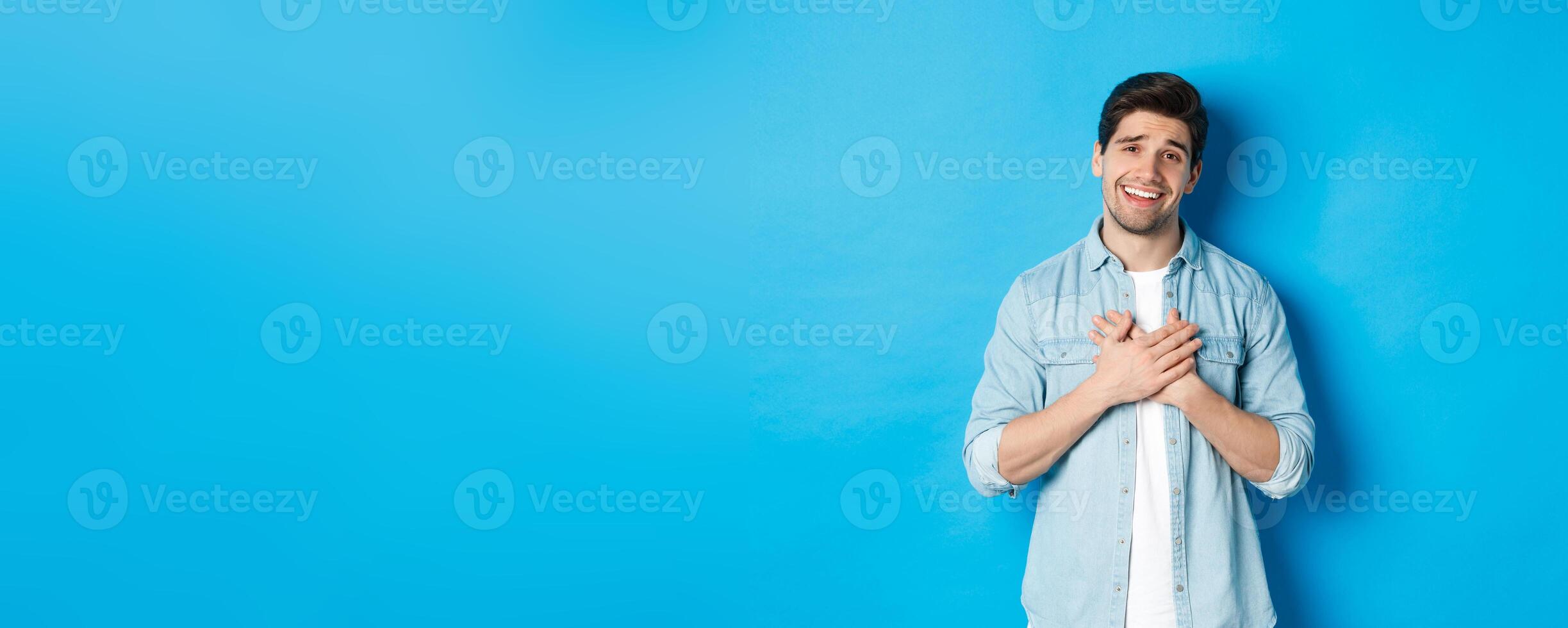 Portrait of flattered handsome man thanking you, holding hands on heart and smiling, looking grateful, standing over blue background photo