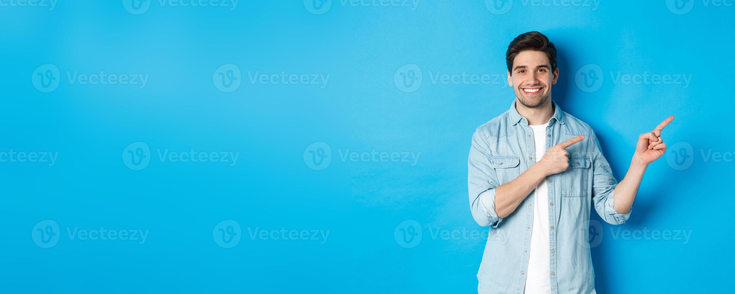 Portrait of attractive adult man smiling, pointing fingers right at logo or banner, showing advertisement against blue background photo