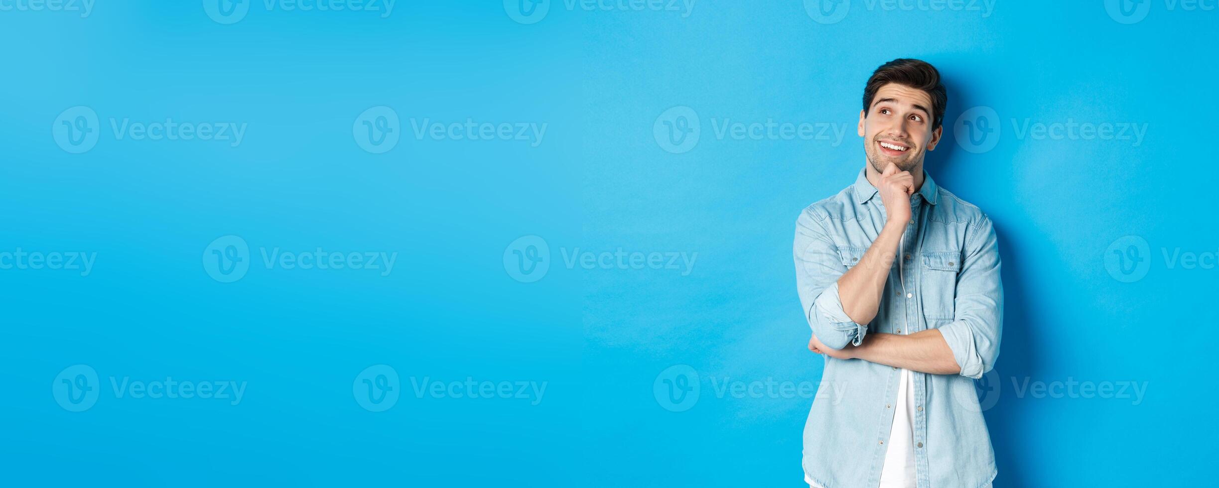 Portrait of thoughtful handsome man with beard, standing in casual outfit, looking at upper left corner and smiling, imaging or dreaming about something, standing over blue background photo
