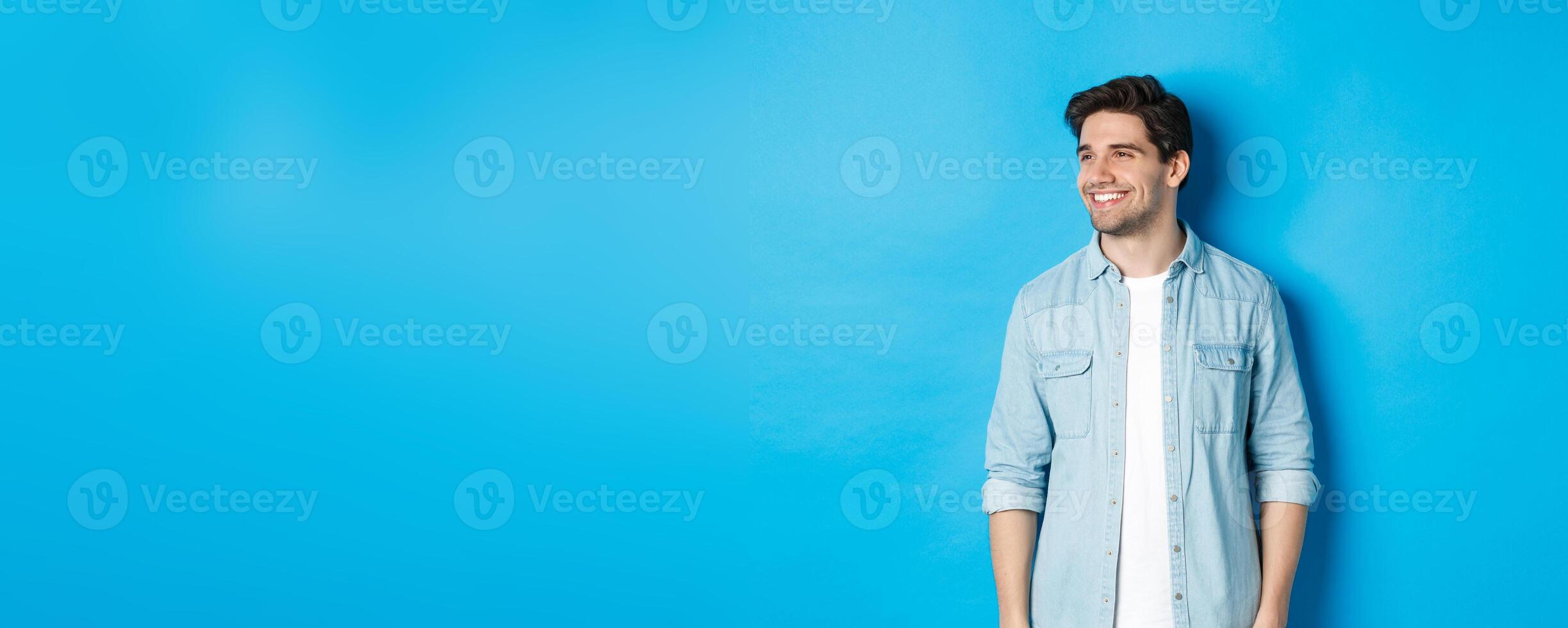 Handsome smiling adult man in casual outfit, smiling and looking left at promo offer, standing against blue background photo