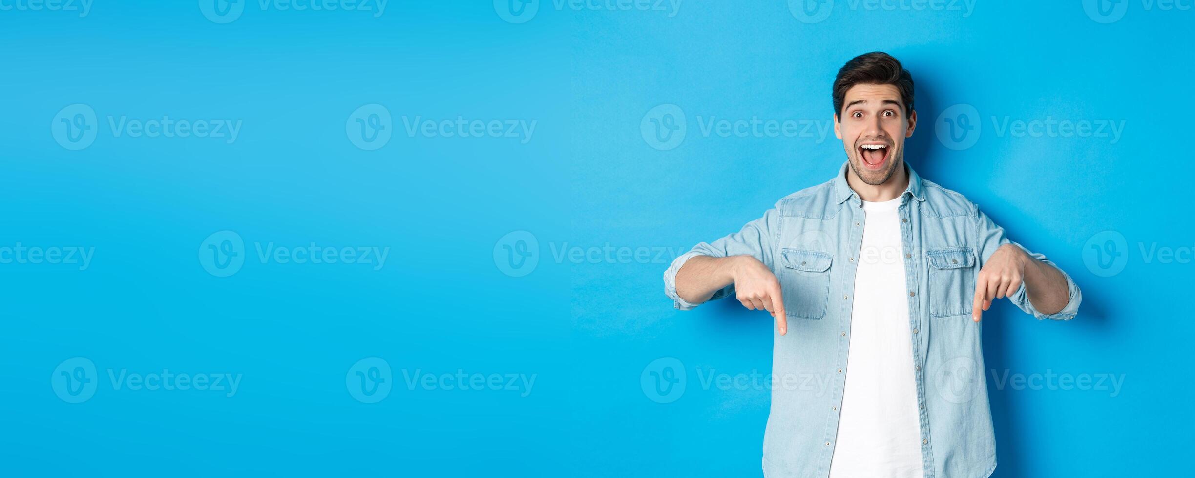 Image of excited handsome man pointing fingers down, making an announcement, standing against blue background photo