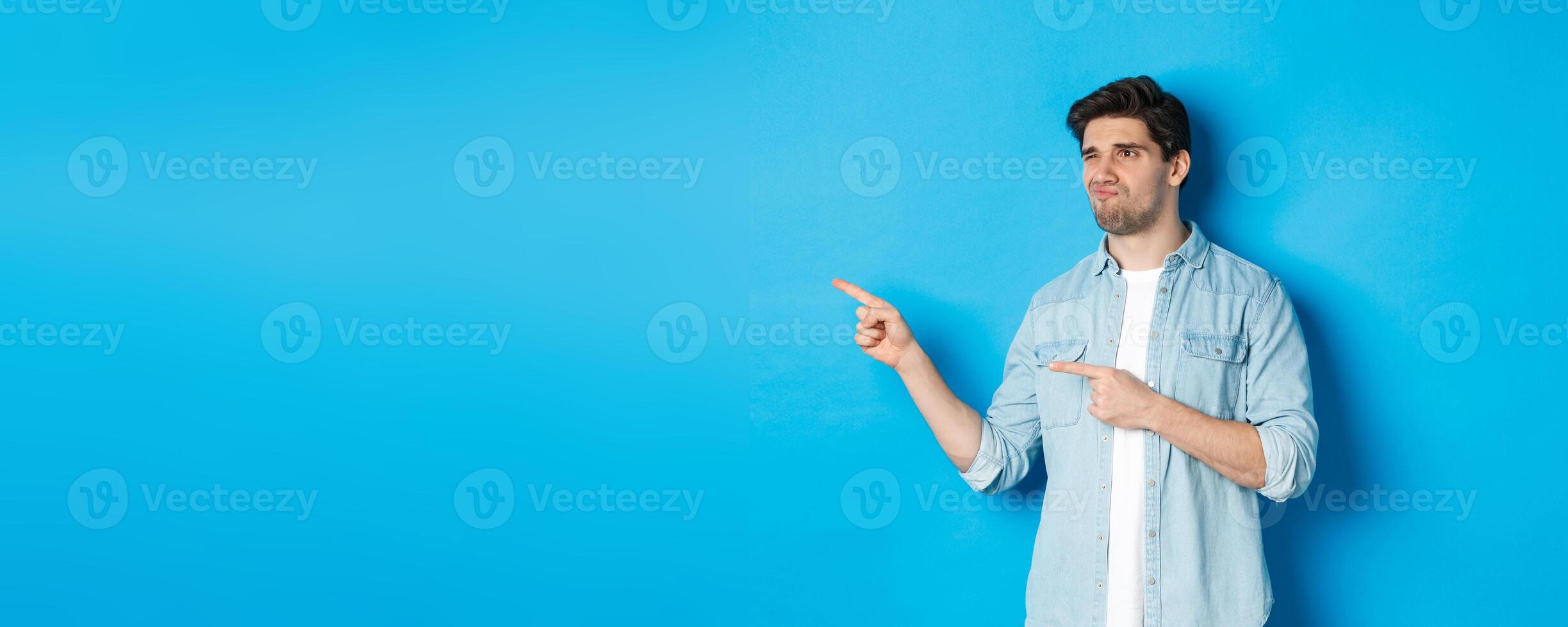 Doubtful adult man pointing fingers left at promotion and looking unsure, grimacing disappointed, standing against blue background photo