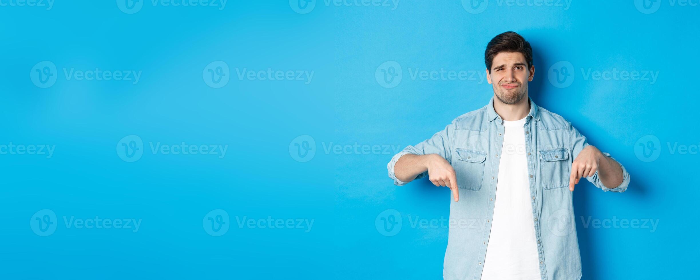 Disappointed guy in casual outfit, pointing fingers down with doubtful grimace, standing against blue background photo