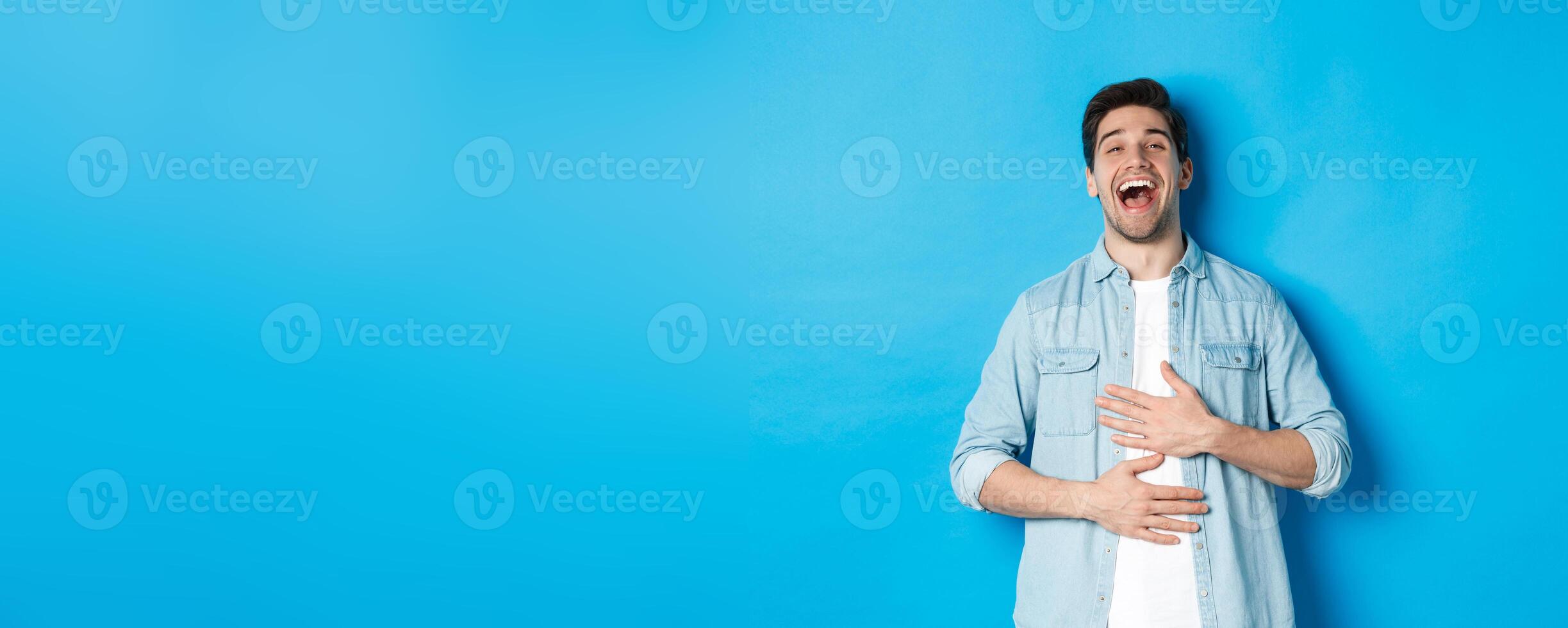 Happy handsome 30 year old man touching belly, laughing out loud and looking at something funny, standing against blue background photo