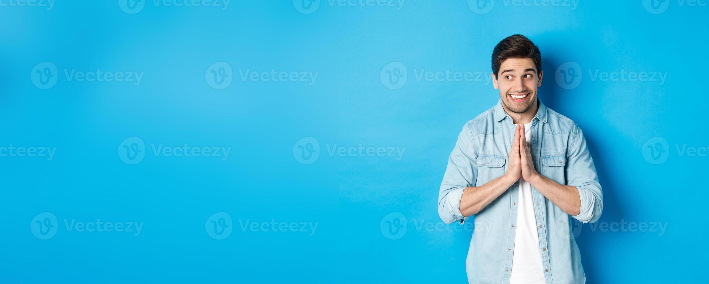 Hopeful smiling man looking left, holding hands in pray, begging for something or pleading, standing against blue background photo