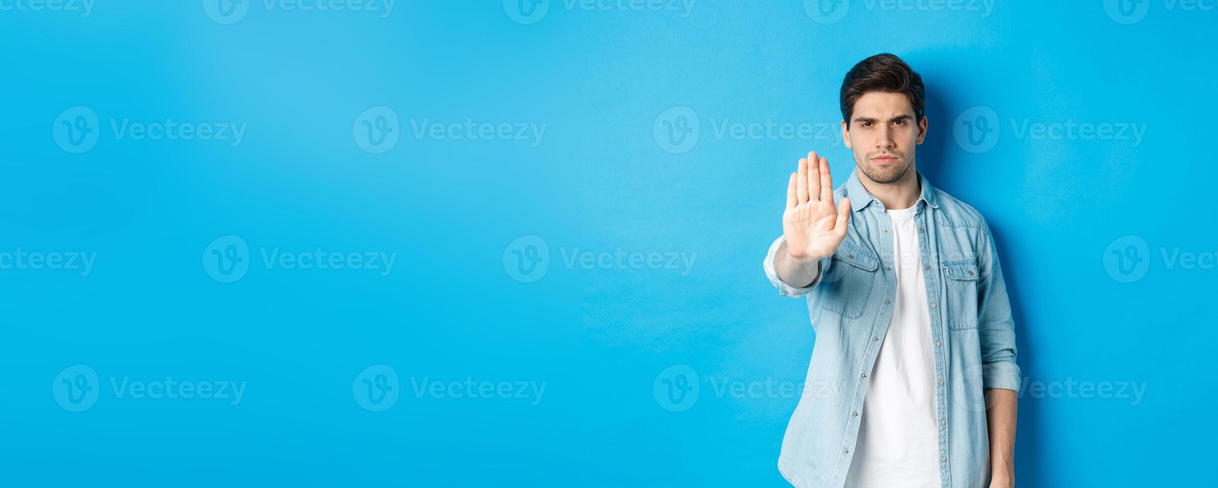 Serious man frowning and saying no, extending hand to shop stop sign, prohibit action, standing against blue background photo