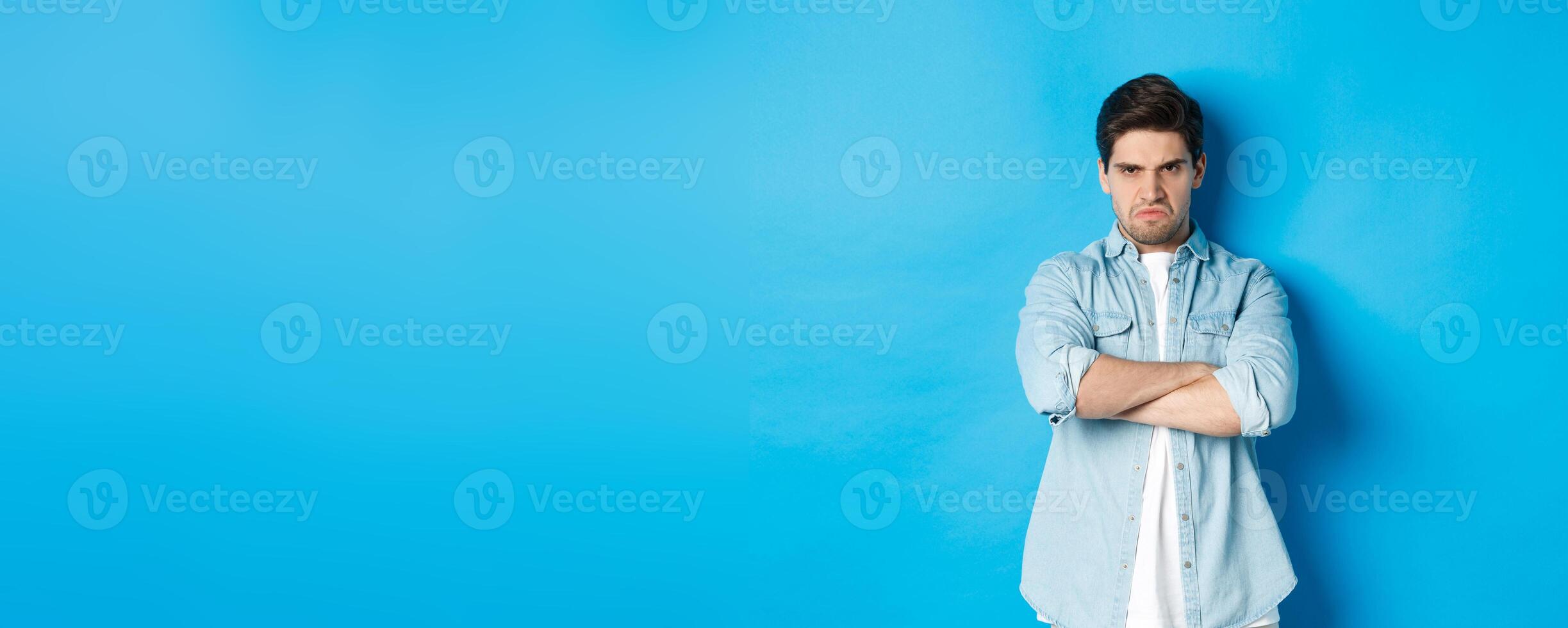 Angry bearded guy crossing arms on chest, frowning, looking mad and offended, standing against blue background photo