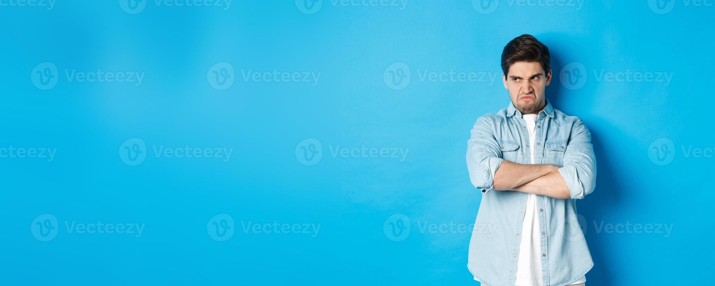 Angry guy cross arms on chest and looking away with insulted expression, standing offended against blue background photo