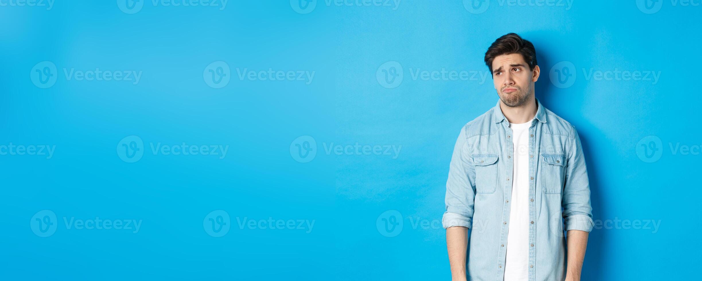 Image of reluctant and sad guy in casual outfit looking left, frowning and feeling upset, standing against blue background photo