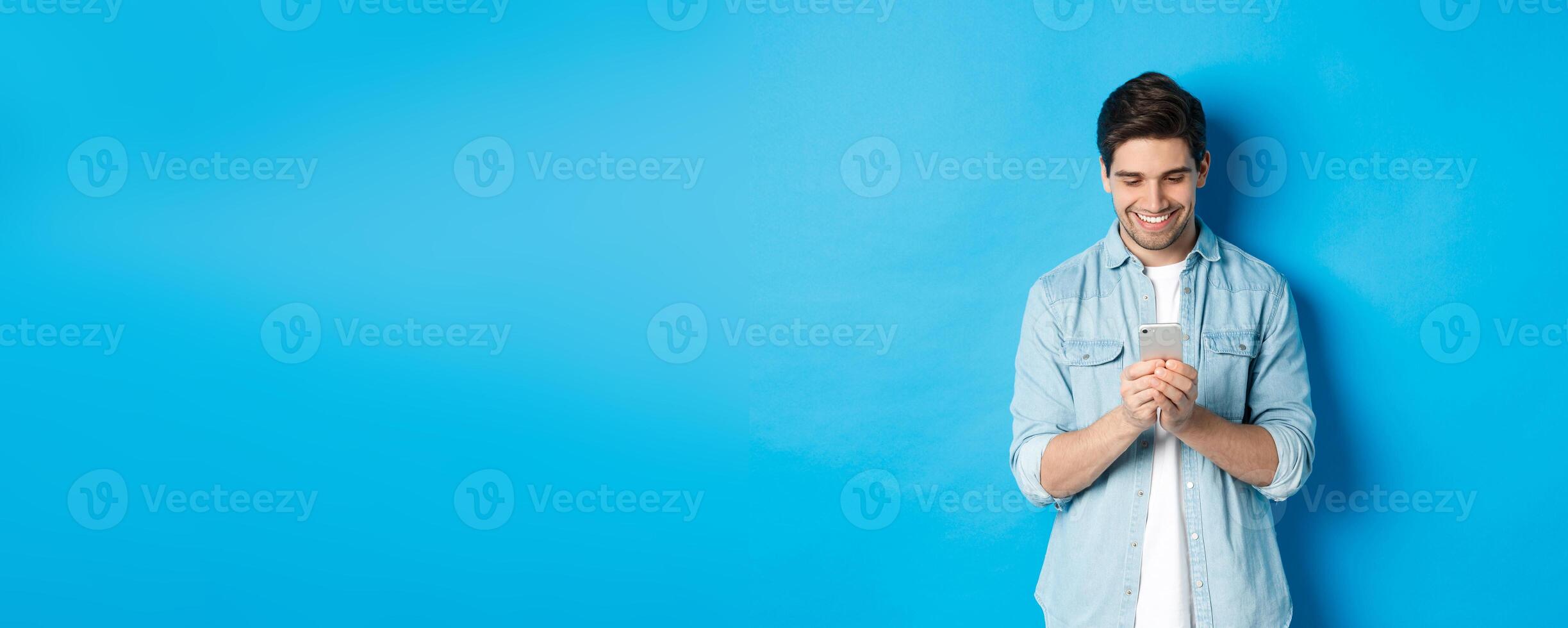 Image of handsome young man using mobile phone, texting on phone and looking pleased, standing over blue background photo