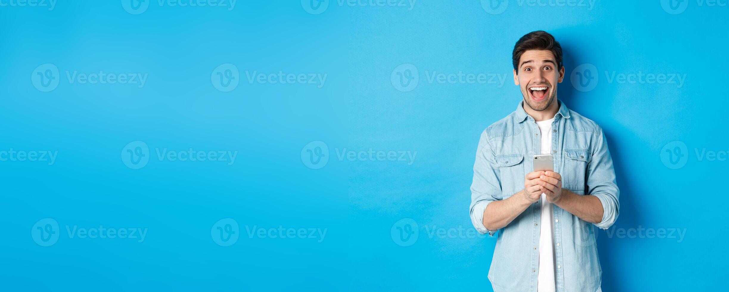 Surprised and happy man winning something online, holding smartphone and rejoicing, standing against blue background photo