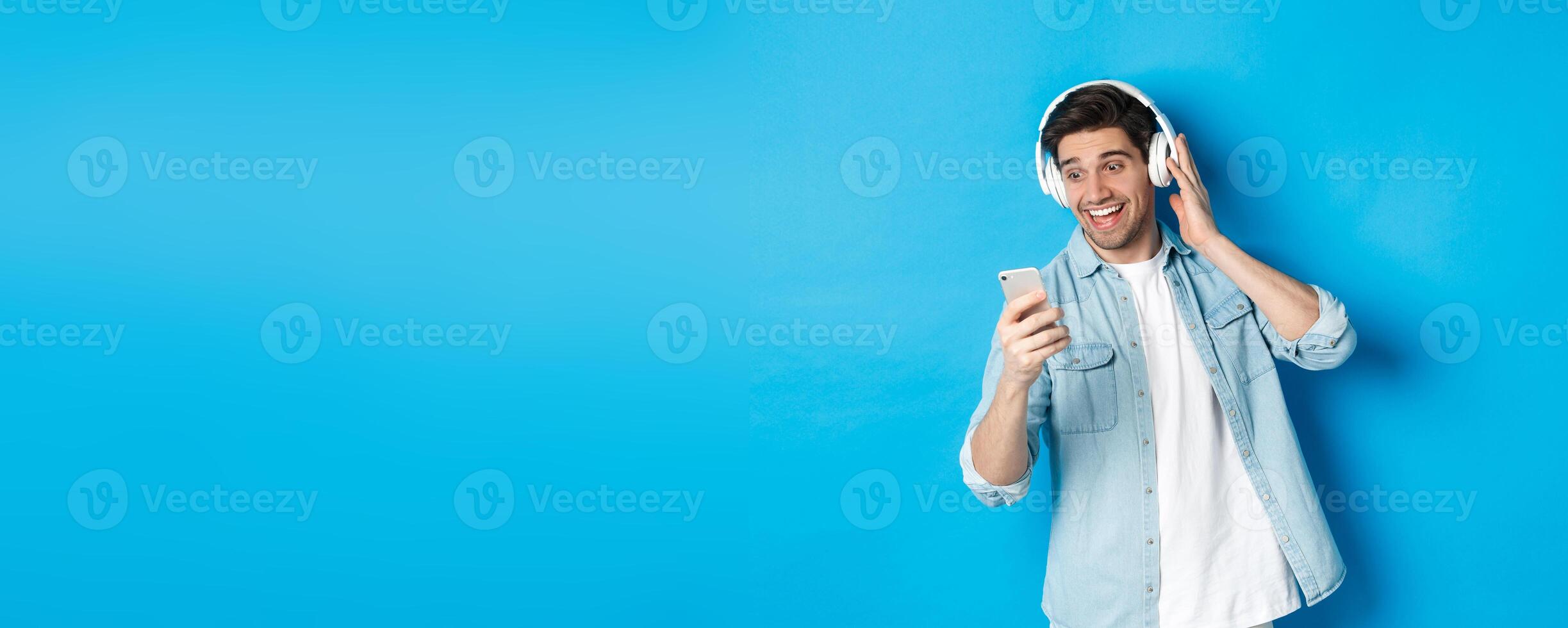 Happy man listening to music in headphones and reading message on smartphone, smiling excited, standing against blue background photo