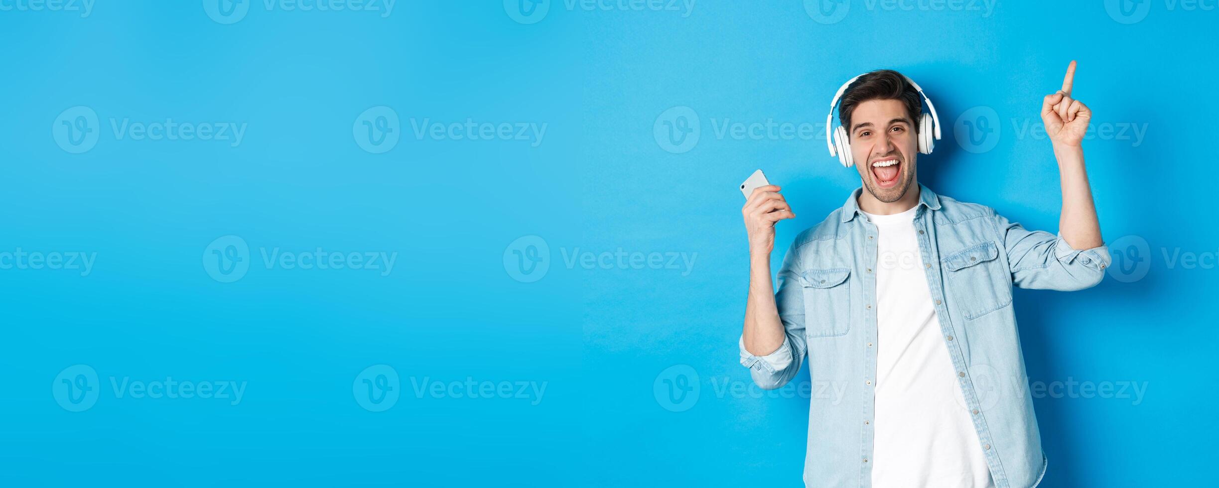 Joyful handsome man dancing with smartphone, listening music in headphones and pointing finger up, standing over blue background photo