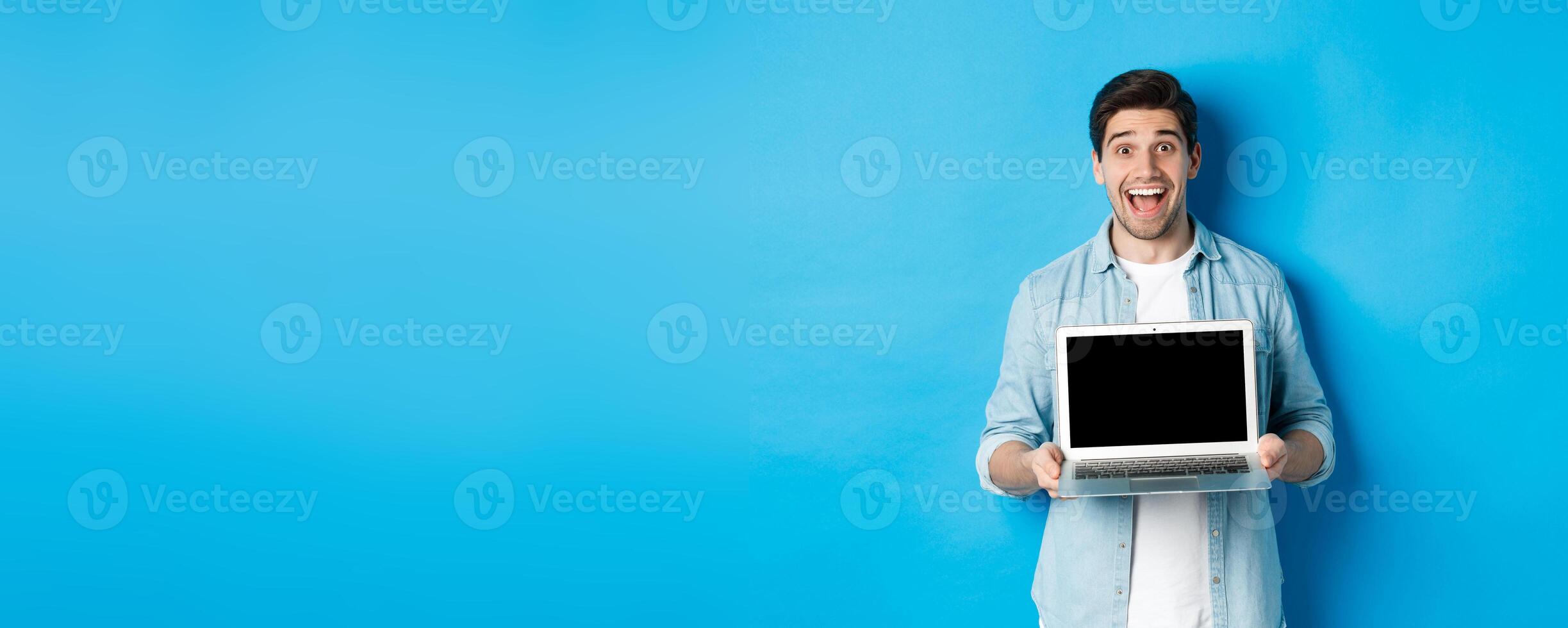 Cheerful smiling man making presentation, showing laptop screen and looking happy, standing over blue background photo